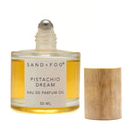 A 50 ml bottle of Sand + Fog Pistachio Dream Eau de Parfum Oil next to its light wood cap, on a white background.