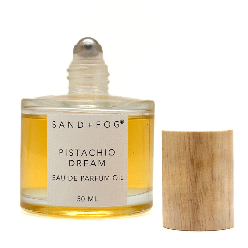A 50 ml bottle of Sand + Fog Pistachio Dream Eau de Parfum Oil next to its light wood cap, on a white background.