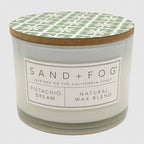 A candle jar with a white wax body, a light wood lid, and a green and white geometric patterned top label. The front label reads SAND + FOG, PISTACHIO DREAM, and NATURAL WAX BLEND.