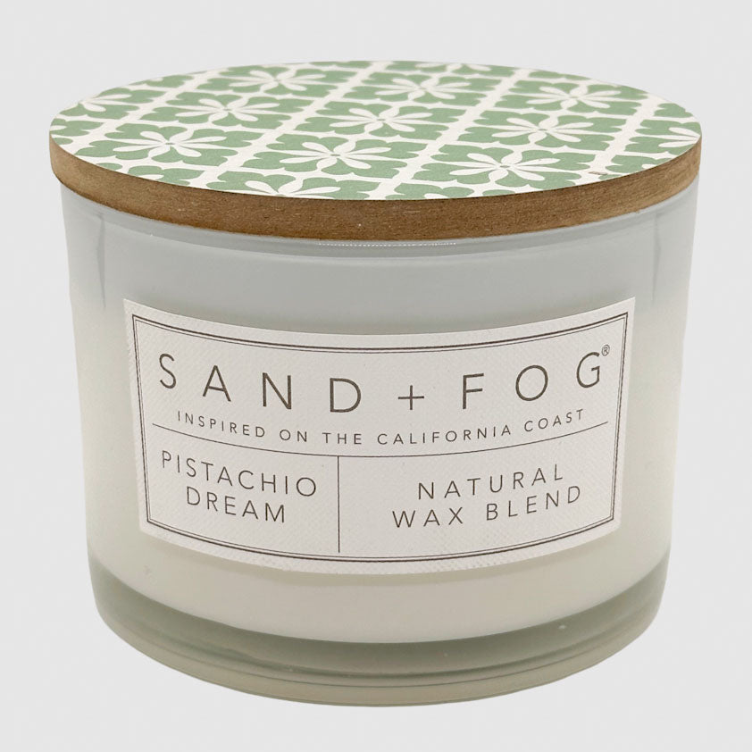 A candle jar with a white wax body, a light wood lid, and a green and white geometric patterned top label. The front label reads SAND + FOG, PISTACHIO DREAM, and NATURAL WAX BLEND.