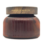 A ribbed DW Home candle in an amber glass jar with a dark brown lid featuring gold foil text that reads 'PUMPKIN BRULEE'.
