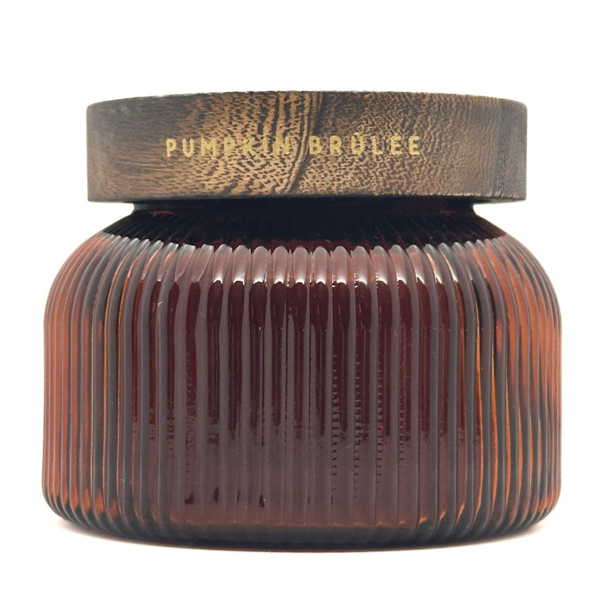 A ribbed DW Home candle in an amber glass jar with a dark brown lid featuring gold foil text that reads 'PUMPKIN BRULEE'.