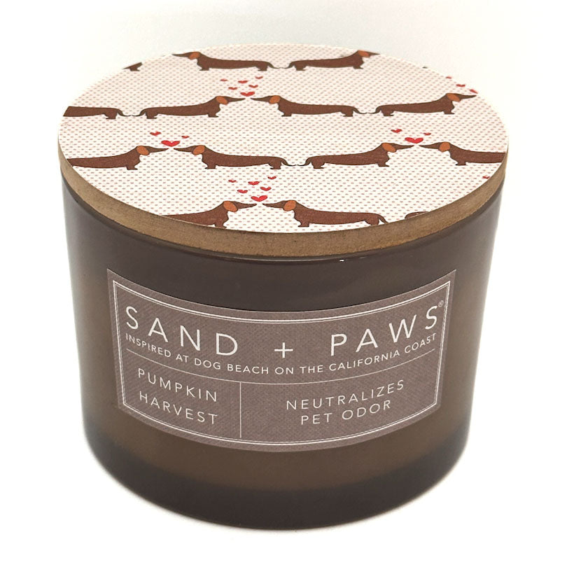 A dark brown candle jar with a lid patterned with dachshunds and small red hearts, labeled 'SAND + PAWS' and 'Pumpkin Harvest Neutralises Pet Odour'.