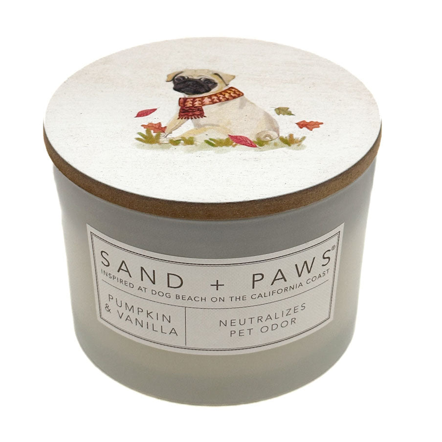 A jar candle with a light grey body and a white lid decorated with an illustration of a pug wearing a red scarf, surrounded by small leaves. The front label reads 'SAND + PAWS' and 'Pumpkin & Vanilla'.