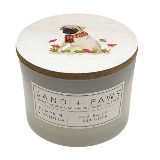 A jar candle with a light grey body and a white lid decorated with an illustration of a pug wearing a red scarf, surrounded by small leaves. The front label reads 'SAND + PAWS' and 'Pumpkin & Vanilla'.