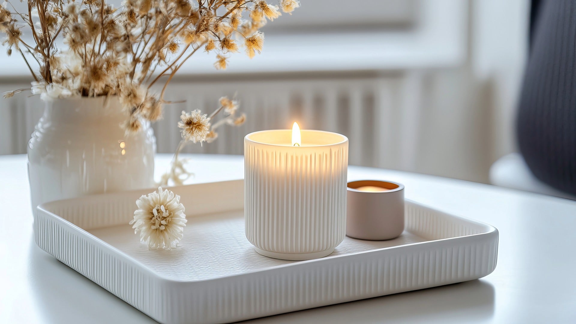 A lit white ribbed candle holder and a small matching candle sit on a white tray on a table, with dried flowers in a vase in the background.