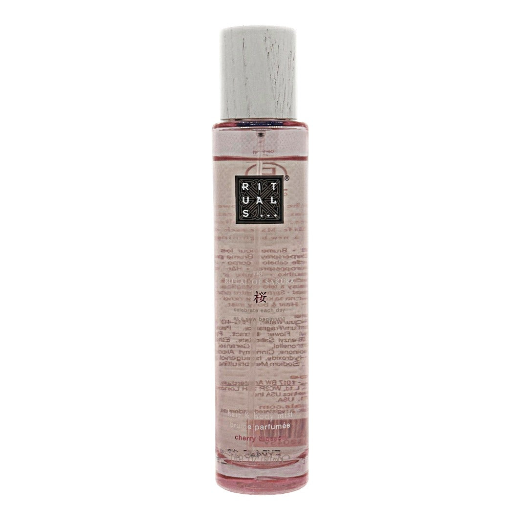 A clear cylindrical bottle of The Ritual of Sakura cherry blossom hair & body mist, containing a pink liquid, featuring a white cap and a black label with white text that reads 'RITUALS'. The bottle is centered against a plain white background.