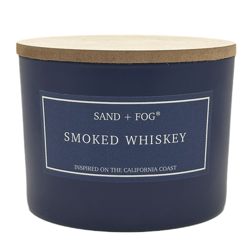 Navy blue candle with a wooden lid labeled 'Sand + Fog Smoked Whiskey' on a white background