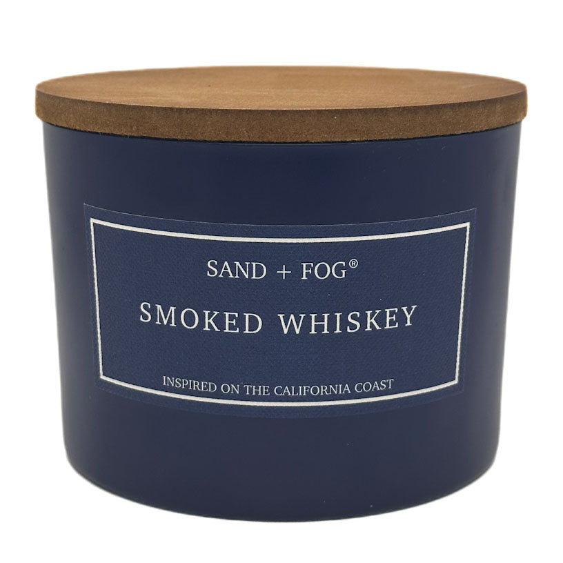 Navy blue candle with a wooden lid labeled 'Sand + Fog Smoked Whiskey' on a white background