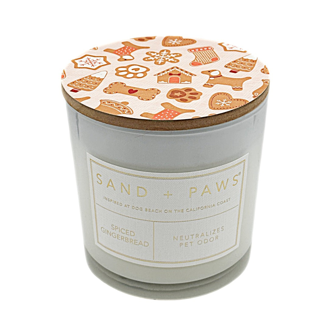 A jar nude candle  and a lid decorated with gingerbread-themed illustrations. The front label reads 'SAND + PAWS' and 'Spiced Gingerbread Neutralises Pet Odour'.