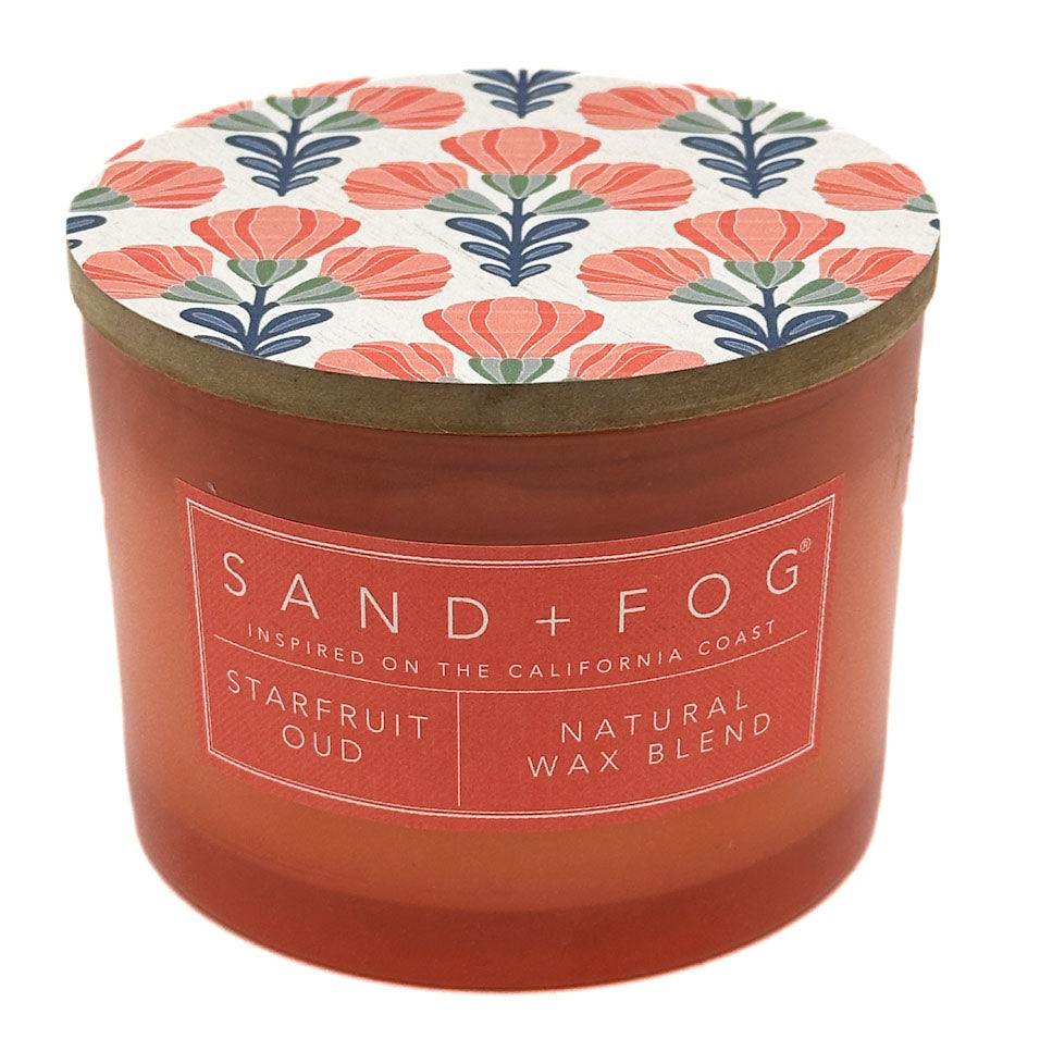 A Sand + Fog Starfruit Oud natural wax blend candle in a translucent terracotta-colored jar with a lid featuring a coral, navy, and green floral pattern on a white background.