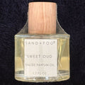 SAND + FOG Sweet Oud perfume oil bottle with wooden cap on a dark textured background