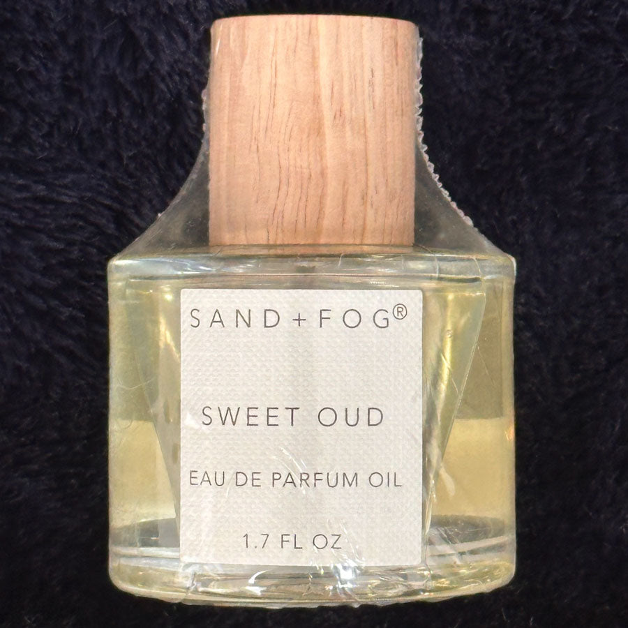 SAND + FOG Sweet Oud perfume oil bottle with wooden cap on a dark textured background