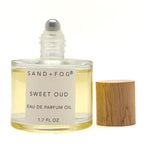 A 50 ml bottle of Sand + Fog Sweet Oud Eau de Parfum Oil next to its light wood cap, on a white background.