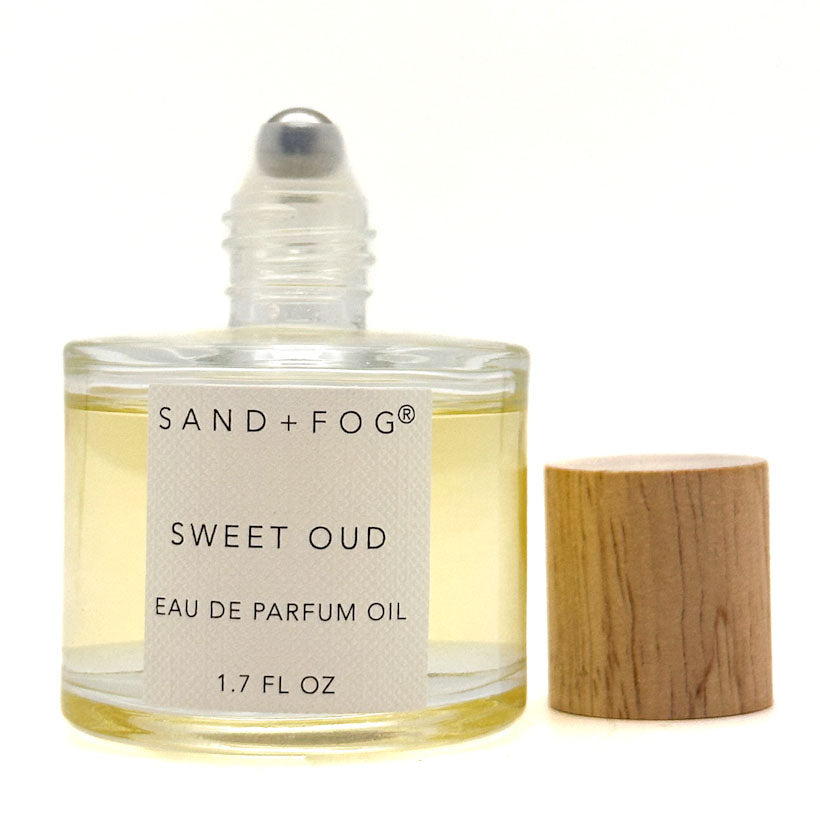 A 50 ml bottle of Sand + Fog Sweet Oud Eau de Parfum Oil next to its light wood cap, on a white background.