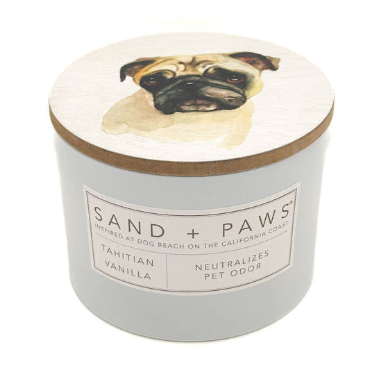 A round jar candle with a light grey body and a wooden lid featuring a painted pug dog illustration. The front label reads 'SAND + PAWS' and 'Tahitian Vanilla Neutralizes Pet Odor'.