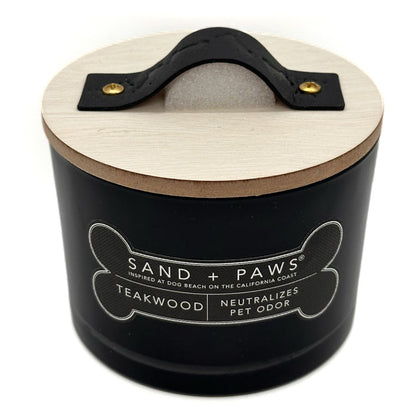 A black cylindrical pet odour neutralirer candle with a light wood lid and a black leather handle. The front label reads 'SAND + PAWS' and 'TEAKWOOD NEUTRALIZES PET ODOR'.