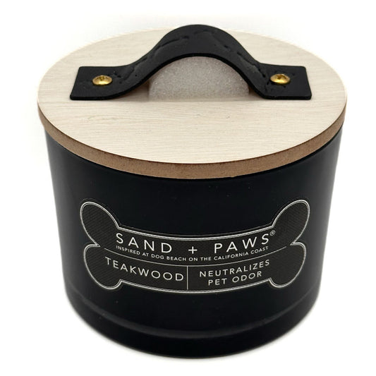 A black cylindrical pet odour neutralirer candle with a light wood lid and a black leather handle. The front label reads 'SAND + PAWS' and 'TEAKWOOD NEUTRALIZES PET ODOR'.