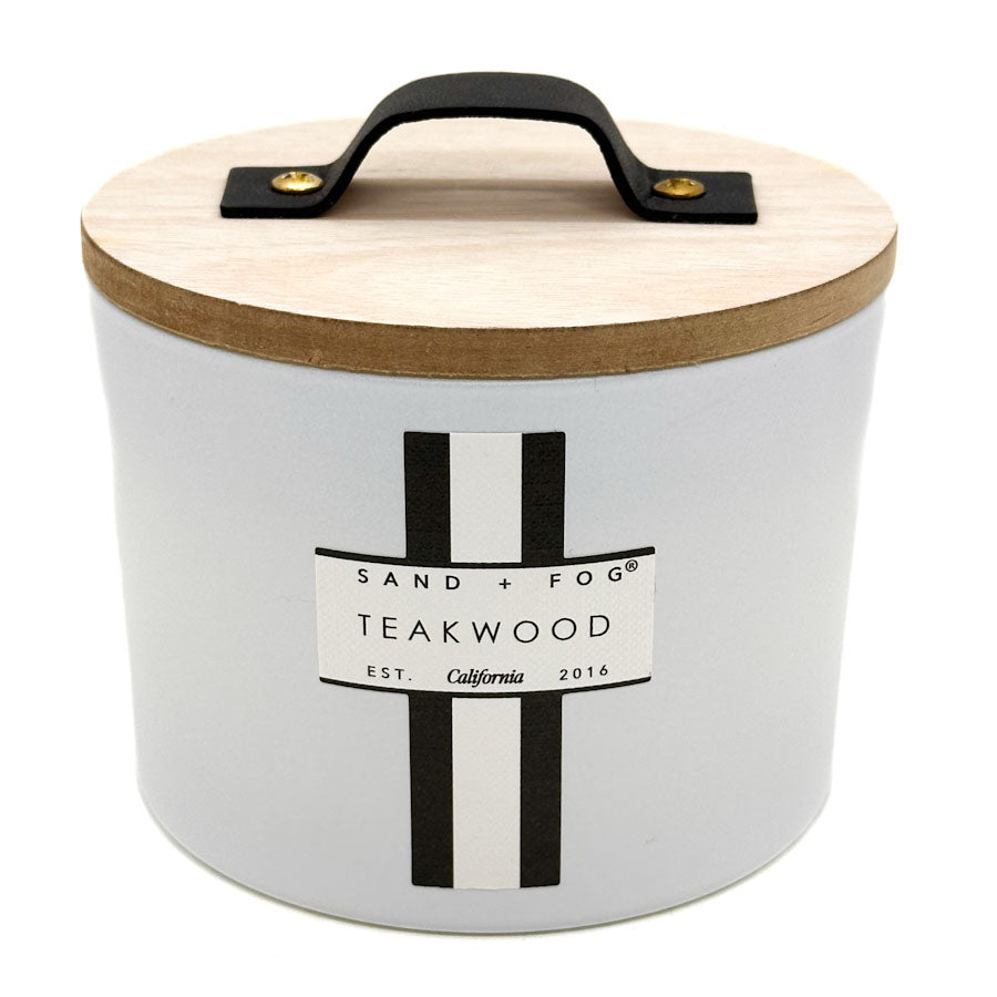 A white cylindrical candle Jar with a light wood lid, a black metal handle, and a black-and-white label reading SAND + FOG TEAKWOOD.