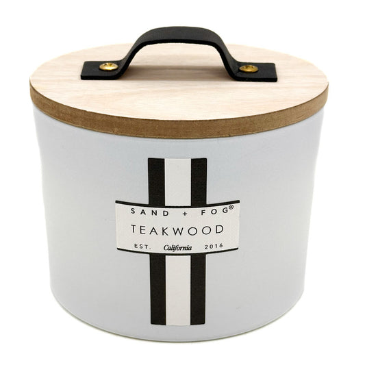 A white cylindrical candle Jar with a light wood lid, a black metal handle, and a black-and-white label reading SAND + FOG TEAKWOOD.