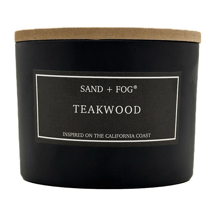 A black jar candle with a light brown kraft paper lid and a black label reading 'SAND + FOG TEAKWOOD' on a white background.