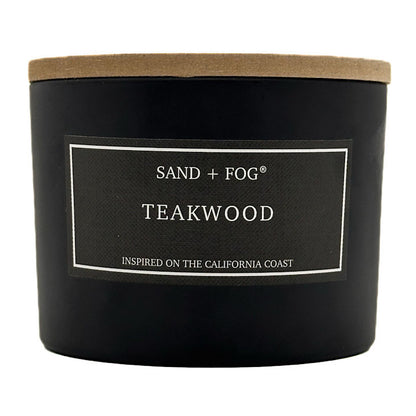 A black jar candle with a light brown kraft paper lid and a black label reading 'SAND + FOG TEAKWOOD' on a white background.