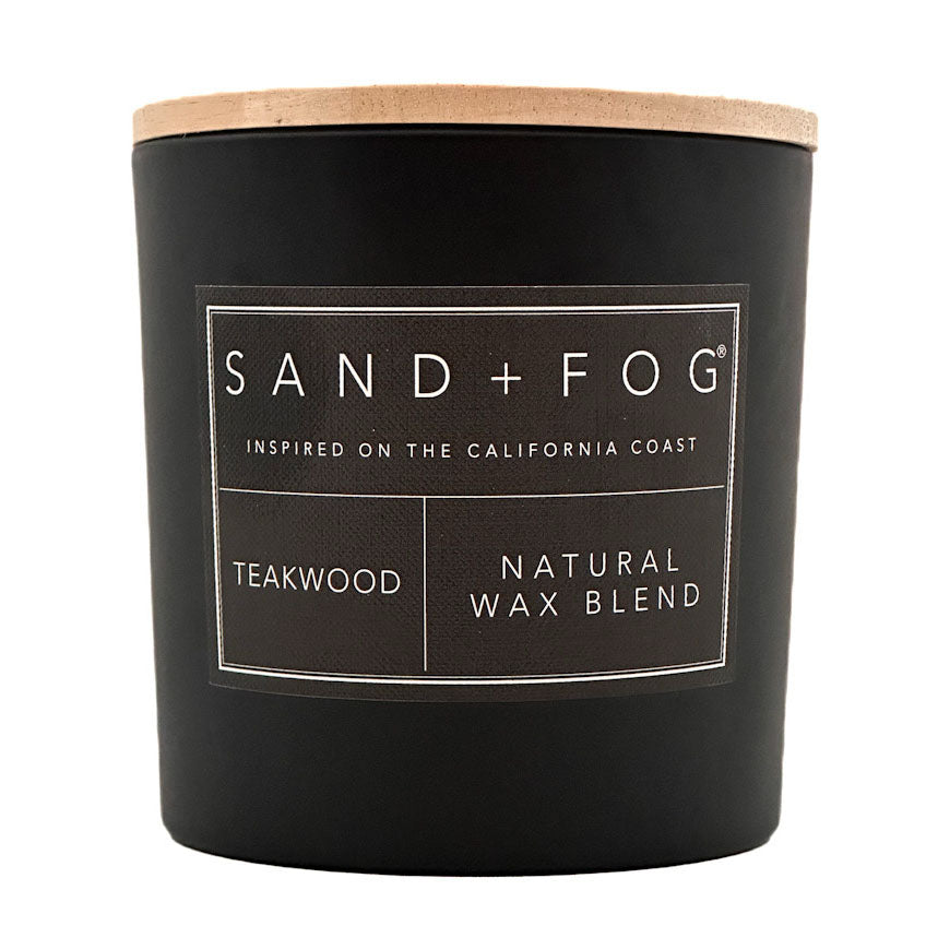 A matte black candle jar with a light natural wood lid and a white printed label reading SAND + FOG, TEAKWOOD, and NATURAL WAX BLEND on a white background.