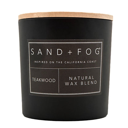 A matte black candle jar with a light natural wood lid and a white printed label reading SAND + FOG, TEAKWOOD, and NATURAL WAX BLEND on a white background.