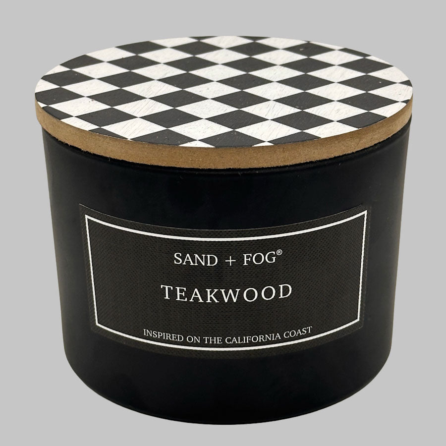A black cylindrical candle jar with a black-and-white diamond-patterned lid and a front label reading 'SAND + FOG TEAKWOOD' on a light gray background.