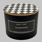 A black cylindrical candle jar with a black-and-white diamond-patterned lid and a front label reading 'SAND + FOG TEAKWOOD' on a light gray background.
