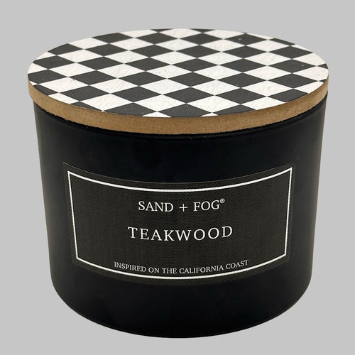 A black cylindrical candle jar with a black-and-white diamond-patterned lid and a front label reading 'SAND + FOG TEAKWOOD' on a light gray background.