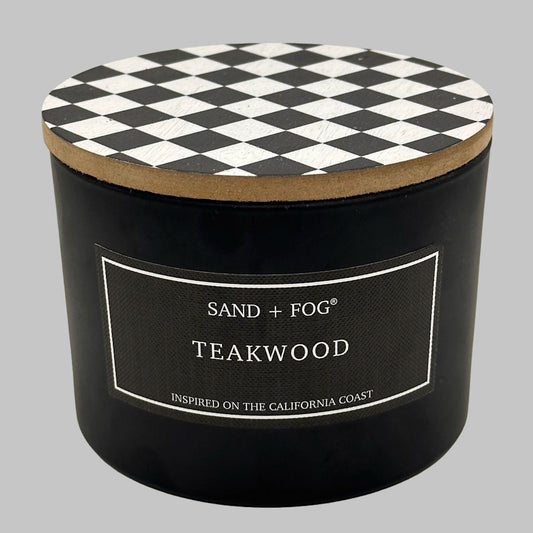 A black cylindrical candle jar with a black-and-white diamond-patterned lid and a front label reading 'SAND + FOG TEAKWOOD' on a light gray background.