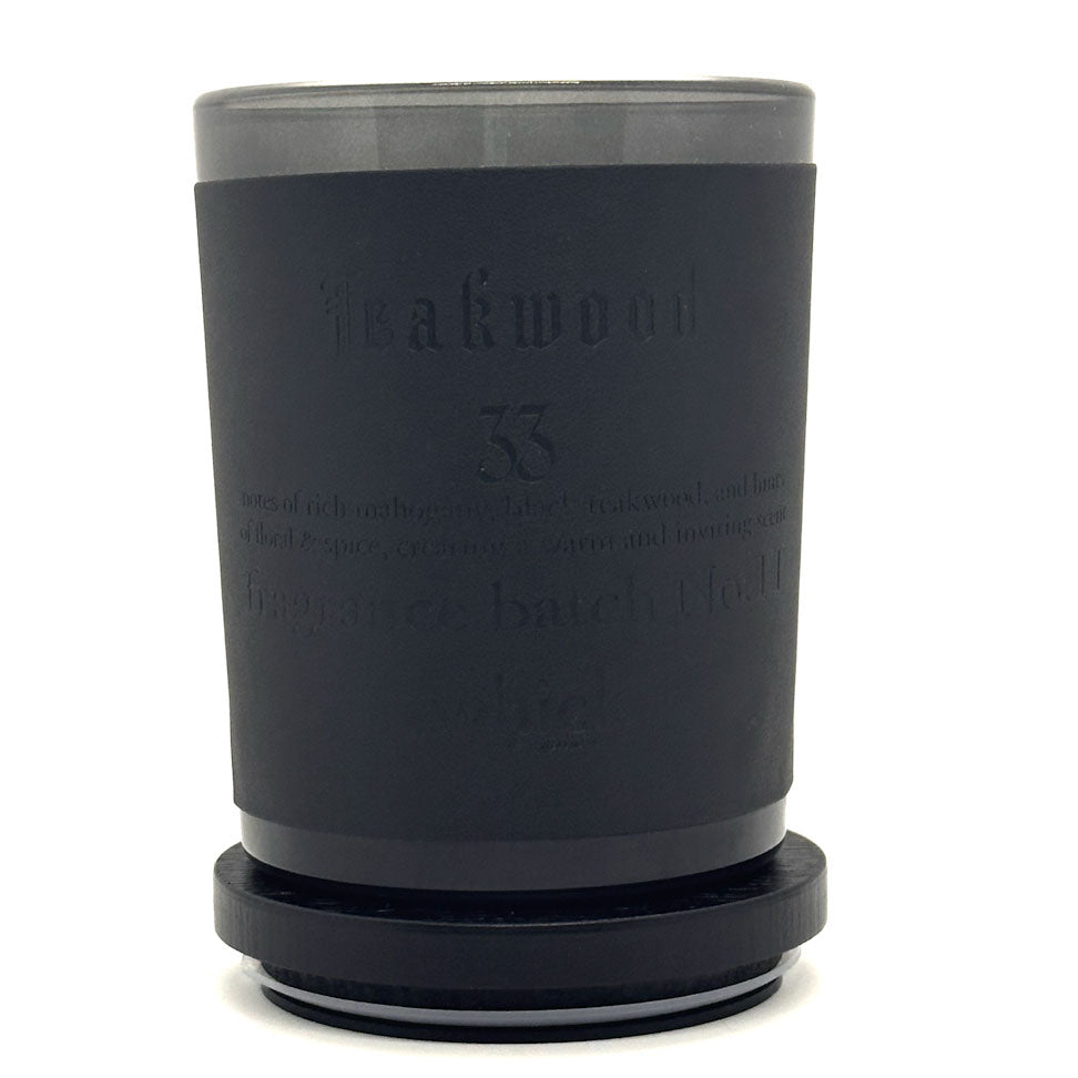 A matte black scented candle in a cylindrical glass container with a matching black lid, featuring embossed text on the front reading 'Teakwood 33'.