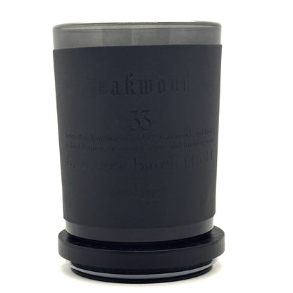A matte black scented candle in a cylindrical glass container with a matching black lid, featuring embossed text on the front reading 'Teakwood 33'.