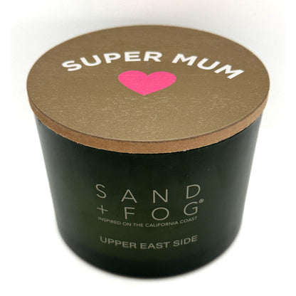 A green cylindrical candle jar with a brown lid, featuring a pink heart and the text 'SUPER MUM' on top, with 'SAND + FOG' Uper East Side printed on the front.