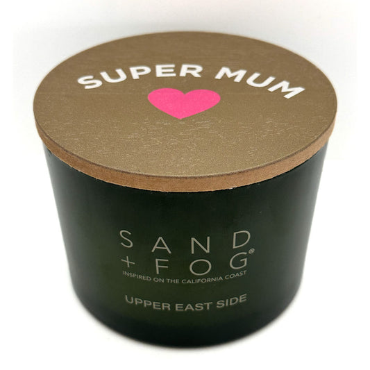 A green cylindrical candle jar with a brown lid, featuring a pink heart and the text 'SUPER MUM' on top, with 'SAND + FOG' Uper East Side printed on the front.