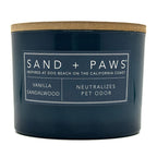 Vanilla Sandalwood Navy blue candle with a wooden lid labeled 'Sand + Paws' on a white background