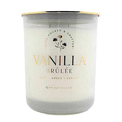 A glass jar candle with white wax and a gold-coloured metal rim, labeled 'VANILLA BRÚLÉE' by DW Naturals, on a solid white background.