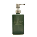 A dark green rectangular pump bottle with a silver metal dispenser, labeled 'SAND+FOG VANILLA SANDALWOOD', on a plain white background.