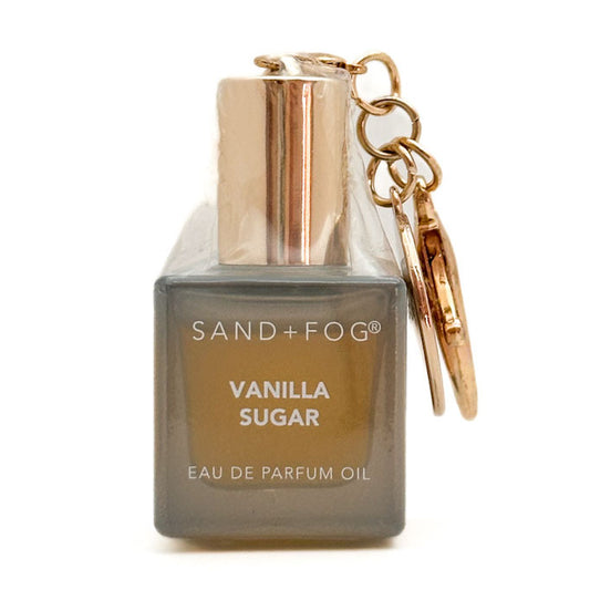 A Sand + Fog Vanilla Sugar Eau de Parfum Oil bottle bag charm with a metallic rose gold cap and matching keychain, set against a plain white background.