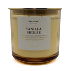 A DW Home Vanilla Brûlée scented candle in an amber glass jar with a gold metal rim and a white paper label, set against a plain white background.