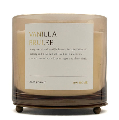 A candle in a clear glass jar with a beige wax fill and a white label reading 'VANILLA BRULEE' and 'DW HOME'. The jar sits on a dark brown wooden stand against a plain white background.