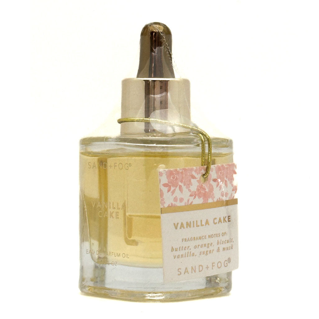 A bottle of Sand + Fog Vanilla Cake Perfume oil with a clear glass body, pale yellow liquid, a metallic dropper cap, and a pink floral label.