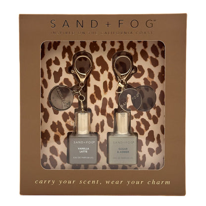 Sand + Fog gift set containing two 10ml travel perfume oil bottles (Vanilla Latte and Sugar & Amber) with leopard print keychains, presented in a brown cardboard box.