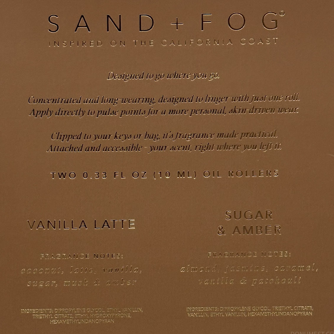 A close-up of a brown cardboard product label for Sand + Fog fragrance oil, showing text for Vanilla Latte and Sugar & Amber scents.