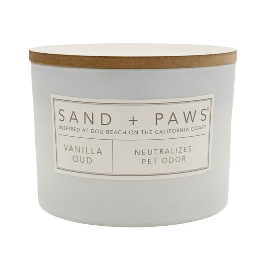 A white jar candle with a light wood lid and a white label reading 'SAND + PAWS' and 'VANILLA OUD'.