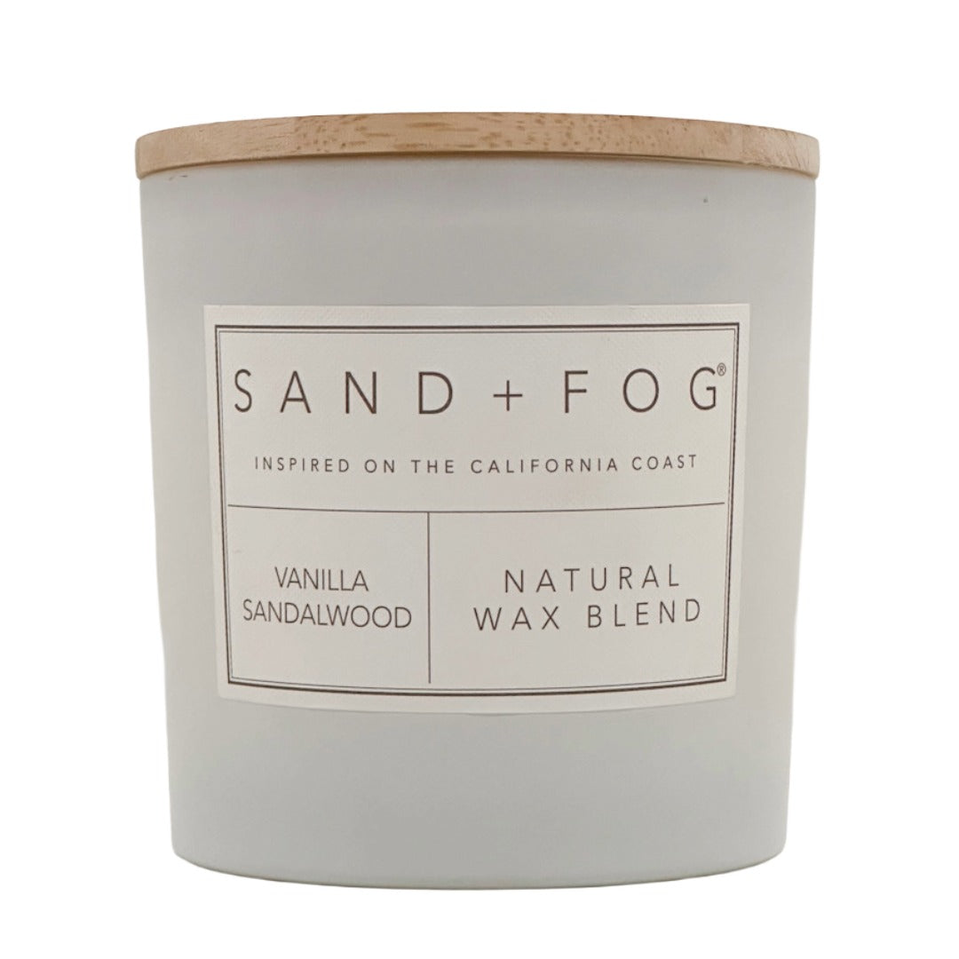 A Sand + Fog candle in a matte light gray jar with a natural wood lid, featuring a white label with black text that reads 'SAND + FOG', 'VANILLA SANDALWOOD', and 'NATURAL WAX BLEND'.