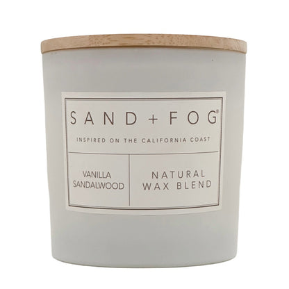 A Sand + Fog candle in a matte light gray jar with a natural wood lid, featuring a white label with black text that reads 'SAND + FOG', 'VANILLA SANDALWOOD', and 'NATURAL WAX BLEND'.