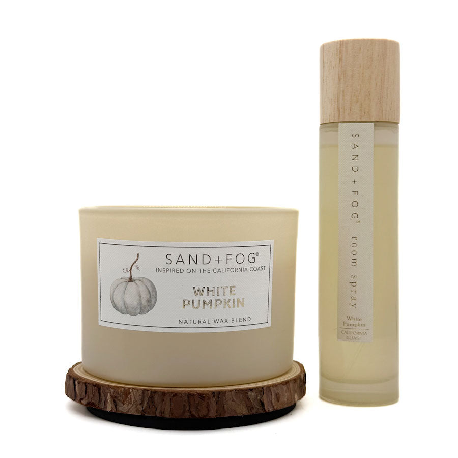 A candle and a room spray set from Sand + Fog in the White Pumpkin scent. The set includes a natural wax candle in a glass jar with a rustic wood slice base, and a clear glass room spray bottle with a light wood cap, both on a white background.