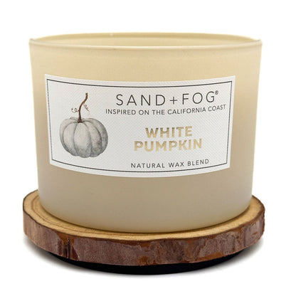 A Sand + Fog 'White Pumpkin' natural wax candle in a cream-coloured glass jar, resting on a round wooden slice coaster against a white background.